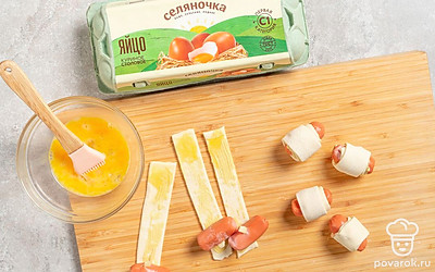 Взбейте куриное яйцо в миске. Немного смазывайте края теста яйцом и скрепляйте их вокруг сосиски. Выложите заготовки на противень, смажьте их яйцом сверху. 