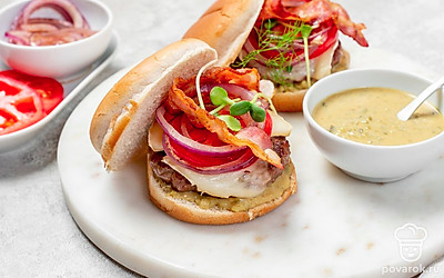 Сделайте соус и соберите бургеры. Смешайте оливковое масло с измельченными корнишонами и горчицей. Нижние части булочек щедро промажьте соусом. Выложите стейки, сверху — ломтики помидоров, кольца лука и бекон. Накройте верхней частью булочки.