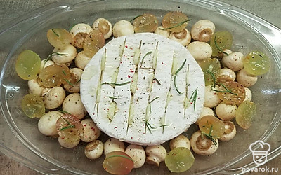 На головке сыра сделайте надрезы, вложите в них пластинки чеснока. Поместите сыр в центр формы для запекания, вокруг разложите ягоды винограда, разрезанные пополам, и шампиньоны. Поперчите, полейте медом и посыпьте розмарином. Поставьте в разогретую духовку и запекайте 15 минут.