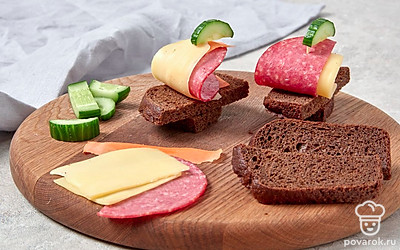 Приготовьте закуску. Возьмите 1 кусочек хлеба и выложите на него ломтики колбасы и огурца. В получившийся бутерброд воткните шпажку. Затем возьмите ломтики сыра и наденьте их на шпажку так, чтобы получились «паруса». На кончик шпажки наденьте треугольник из морковки. Повторите действия со всеми ингредиентами.