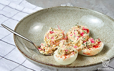 Сделайте начинку. Крабовые палочки измельчите вручную или с помощью терки, отложив одну для украшения. Укроп мелко нарежьте и добавьте ингредиенты в миску с желтками. Посолите по вкусу, добавьте йогурт и смешайте компоненты до однородности. Готовую начинку распределите по половинкам белков.