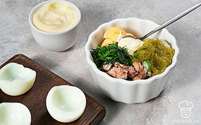 Приготовьте фаршированные яйца. Смешайте в миске желтки, сайру, корнишон и лук. Добавьте петрушку, укроп, майонез и перемешайте. В каждую половинку белка выложите начинку с горкой.