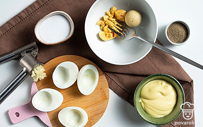 Подготовьте основу. Отваренные яйца очистите от скорлупы, разрежьте на половинки, разложите их на плоском блюде. Желтки извлеките и разомните вилкой в отдельной емкости.