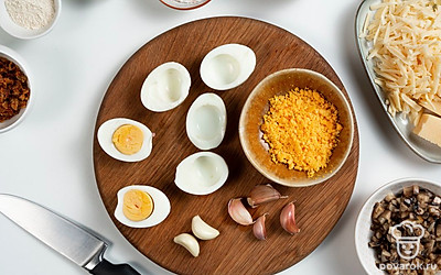 Подготовьте ингредиенты для начинки яиц. Лук нарежьте мелким кубиком. Брызните масло в сковороду и выложите лук. Жарьте до золотистого цвета. Грибы измельчите. Добавьте их к луку. Обжарьте до выпаривания жидкости. Яйца разрежьте пополам. Желтки разомните вилкой. Сыр натрите на крупной терке. Петрушку накрошите. У кудрявой петрушки для подачи отделите веточки от стеблей.