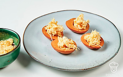 Приготовьте закуску. Желток разомните вилкой, смешайте с сыром, чесноком, майонезом. Зафаршируйте яичные белки начинкой. Поставьте на плоское блюдо срезанным краем вниз. Сверху выложите окрашенные крышечки, чтобы получились грибочки.