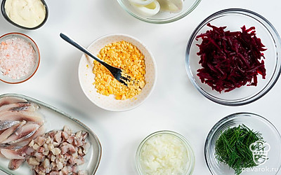 Подготовьте ингредиенты для начинки яиц. Яйца разрежьте пополам. Желтки уберите в отдельную посуду. Свеклу натрите на крупной терке. Лук измельчите. Залейте его уксусом и водой в равных пропорциях, оставьте мариноваться на 15 минут. Чеснок продавите в прессе. Селедку очистите, уберите косточки, разделайте на филе. Одну часть филе нарежьте мелко, другую — ломтиками. Желток накрошите.