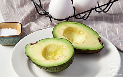 Подготовьте ингредиенты блюда. В емкость, подходящую для микроволновой печи, выложите половинки авокадо отверстиями от косточек вверх. В каждую половинку разбейте по одному яйцу, сохраняя желток целым, и посолите.