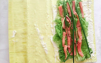 Затем выложите листья салата, заняв примерно треть части листа лаваша. Сверху на салат выложите зеленый лук и кусочки рыбы. Сверните лаваш в плотный рулет и нарежьте на порционные кусочки.