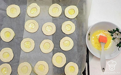 Чтобы тесто хорошо и равномерно поднялось, все заготовки наколите вилкой. В небольшой емкости взбейте яйцо, при помощи вилки. На подготовленный противень с пергаментной бумагой, выложите кружки из теста, смажьте их яйцом, затем сверху выкладывайте кольца и тоже смазываем яйцом.