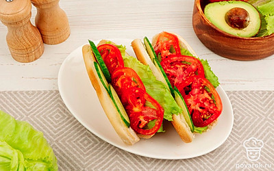 Булочки для хот-догов разрежьте вдоль, но не до конца. Раскройте их и выложите с обеих сторон листья салата, ломтики огурца и помидора.