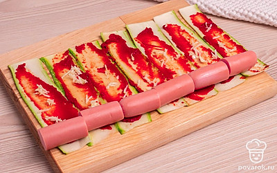 Нарежьте сосиски на несколько частей. Каждую овощную пластинку смажьте томатной пастой, на край положите дольку сосиски. Мелко нарежьте чеснок и добавьте в рулетик.