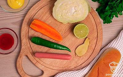 Подготовьте все ингредиенты и выложите их на стол. Снимите пленку с сосисок. Помойте острый перец и петрушку под проточной водой. Помойте и очистите морковь. Снимите верхние листья с кочана капусты.