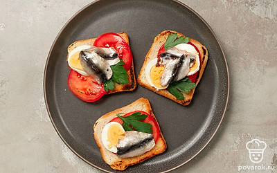 Приготовьте блюдо. Обжаренные хлебные ломтики обильно натрите чесноком с обеих сторон, переложите на большую тарелку. Сверху распределите кружочки помидоров, листья кинзы и ломтики яиц. Наверх выложите филе анчоусов.