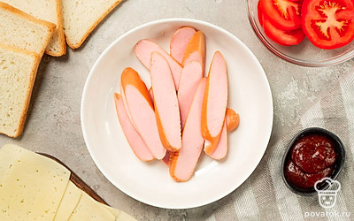 Измельчите продукты. Помидор нарежьте кружочками, сыр — ломтиками, сосиски разрежьте вдоль на полоски.
