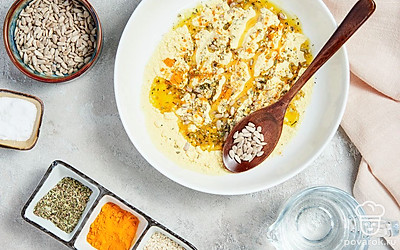 Приготовьте основу для хлебцев. В миску пересыпьте просеянную муку. Добавьте к ней семечки, кунжут, прованские травы, куркуму и соль, хорошо перемешайте. После этого влейте в смесь горячую воду и масло, еще раз тщательно перемешайте. Готовое тесто скатайте в шар, обмотайте его пленкой и оставьте на 20 минут.