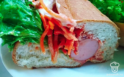 Поместите внутрь булок поджаренную сосиску, полукольца лука, морковь по-корейски, украсьте листьями салата, залейте соусом из оставшегося майонеза и кетчупа.