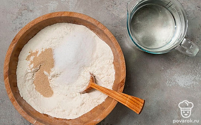 Приступите к замесу теста. Для этого в миску, подходящую для замеса, всыпьте все сыпучие продукты: муку, дрожжи, соль. Влейте подогретую воду, перемешивайте сначала ложкой, затем — рукой. Не вымешивайте. Переложите в другую емкость, предварительно смазанную маслом, прикройте пищевой пленкой и оставьте в тепле на 35-40 минут.