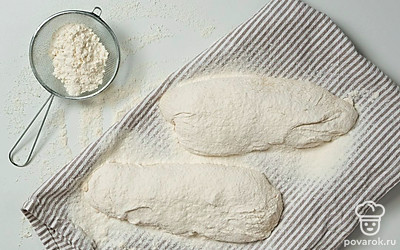 Сформируйте багеты-чиабатты. Щедро присыпьте поверхность стола мукой. Выложите тесто на стол, обваляйте в муке, разделите на две части, сформируйте широкие приземистые батоны. Каждую часть теста переложите на обсыпанное мукой полотенце, зафиксируйте по бокам твердыми предметами, чтобы тесто не растеклось. Оставьте на 20 минут.