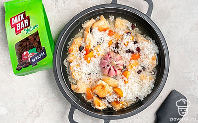 Доведите плов до готовности. Когда содержимое казана закипит, убавьте огонь. Всыпьте изюм Mix Bar и курагу «Красная цена». Перемешайте. Положите в центр головку чеснока Global Village. Накройте казан крышкой и готовьте 20–25 минут.