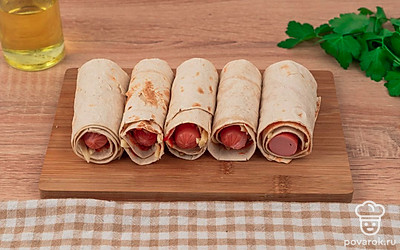 Сыр потрите на крупной терке и присыпьте им смазанные полоски лаваша. Заверните сосиски.