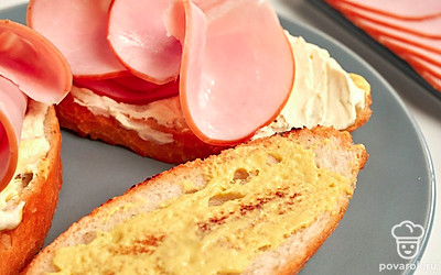 Приготовьте багет с копченой свининой. Копченую свинину нарежьте тонкими ломтиками. Каждый кусочек багета поверх горчицы намажьте плавленым сыром, сверху красиво положите несколько сложенных пополам кусочков копченой свинины.