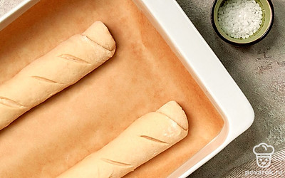 Запеките багет на кислом молоке. Тесто выложите на стол, еще раз хорошо промните руками. Оно должно немного липнуть. Разделите массу на две части. Каждую раскатайте в прямоугольник. Затем сверните в рулет. Края защипните. Выложите на противень с пергаментом швом вниз. Оставьте на 15 минут. Ножницами, ножом или лезвием сделайте сверху надрезы по диагонали. Запекайте при 160-180 градусах 30-40 минут.