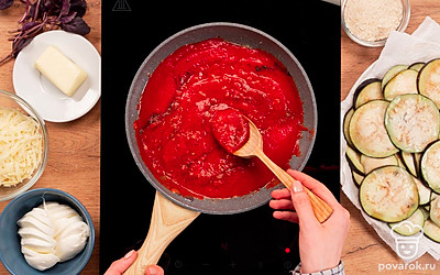 Пока уходит влага из баклажанов, сделайте зажарку. Разогрейте сковороду, влейте масло, выложите мелко нарезанный лук и чеснок. Обжарьте до румяности на среднем огне и добавьте консервированные помидоры в собственном соку. Помидоры немного разомните. Добавьте соль, перец, специи по вкусу, базилик и перемешайте. Доведите до кипения, уменьшите огонь до минимума и тушите 15 минут.