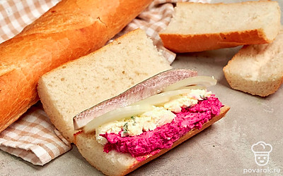 Приготовьте багет с филе сельди. Разрежьте хлеб на две половины. Затем каждую из частей разделите вдоль на две части. Мякиш примните. Выложите начинку слоями: свекольная масса, смесь с зеленью, кольца лука, филе рыбы, маринованные огурцы. Закройте бутерброды вторыми половинами багета. Заверните в пленку, уберите в холод на час.