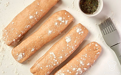 Сформируйте багеты. Присыпьте стол мукой, положите на него тесто и разделите его на 4 части. Из каждой части сформируйте небольшие прямоугольники. Распределите смесь из базилика и сливочного масла по заготовкам из теста. Сверните заготовки в рулеты, сделайте на них вертикальные надрезы. Разложите рулеты из теста на противне, сделав между ними перегородки из пекарской бумаги.