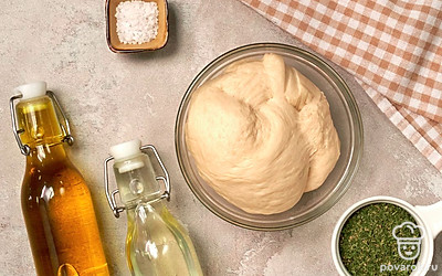 Приготовьте тесто. Возьмите емкость с теплой водой, добавьте соль и сахар. В другой миске смешайте муку и дрожжи. Сделайте воронку в муке и влейте туда смесь из воды, сахара и соли. Хорошо замесите тесто руками до соединения всех ингредиентов. Выложите его в чистую посуду, смазанную растительным маслом. Отправьте емкость с тестом в теплое место, накрыв пищевой пленкой. Через 40 минут снова замесите, накройте и отложите в тепло на 35 минут.