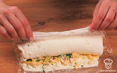 Приступите к оформлению рулета. Сырный пласт освободите от верхнего слоя упаковки. Ровным слоем распределите начинку из белков. Затем нанесите слой начинки с крабовыми палочками. Сверху присыпьте зеленью. С помощью пленки сформируйте сырный рулет и уберите в морозильную камеру на 2 часа.
