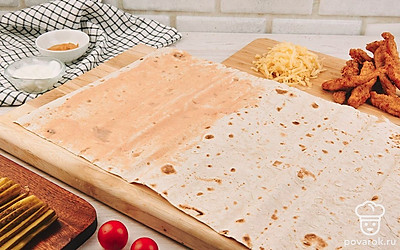 Соберите ролл. Лаваш выложите на плоскую поверхность, половину листа лаваша смажьте соусом, а на другую выложите куриные стрипсы и сыр.