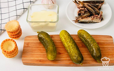 Переложите шпроты из банки в другую емкость и слейте масло. Мелко порубите соленые огурцы и чеснок. Разомните шпроты вилкой до однородной массы из небольших кусочков.