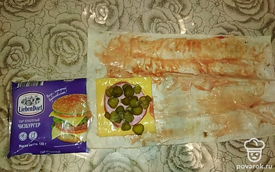 Далее с края выложите ломтик плавленого сыра. Поверх него положите кружок вареной колбасы. Сверху нарежьте кольцами маринованные огурчики. 