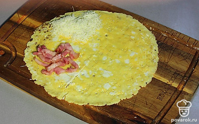 Переложите яичный блин на доску. Сделайте надрез снизу до середины. Положите в левый нижний угол грудинку. Сверху распределите сыр.