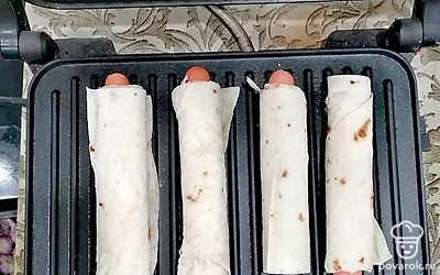 Плотно сверните лаваш. Заверите в лаваш остальные сосиски. Рулетики выложите на гриль, запекайте 7-8 минут.