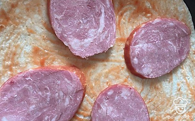 Смажьте основу кетчупом. Нарежьте тонкими ломтиками колбасу и выложите сверху.