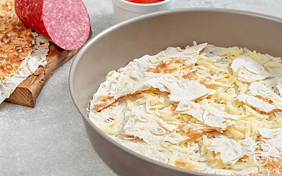 Вырежьте из лаваша круги по размеру формы. Их должно получиться два. Обрезки не выбрасывайте. Форму смажьте маслом или застелите пергаментом. Выложите одну заготовку из лаваша. Треть сыра натрите и присыпьте лаваш. Поверх сыра равномерно выложите часть обрезков от лаваша, покрошив их.