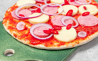 Приготовьте пиццу. Выложите на противень основу для пиццы. Смажьте небольшим количеством оливкового масла, обильно промажьте соусом. Выложите колбасу, кольца лука, сверху моцареллу. Равномерно разложите чили. Поставьте в разогретую до 220 градусов духовку на 15 минут.