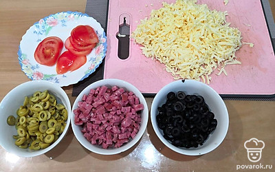 Нарежьте помидоры, оливки и маслины кружочками. Сервелат нарежьте кубиками, сыр натрите на крупной терке.