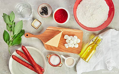 Нагрейте воду до 37-40 градусов. Достаньте моцареллу из рассола и нарежьте тонкими кружочками. Порежьте кружками охотничьи колбаски, маслины нарежьте колечками. Пармезан натрите на мелкой терке и отложите 1-2 ст. л. для подачи. Базилик помойте и разделите на листики. Приготовьте широкий противень и разогрейте духовку до 220-230 градусов.