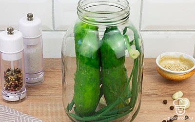 В заготовленную банку положите чеснок, пучок зеленого лука. Вертикально заложите в банку огурцы.