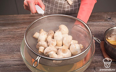 Отварите грибы. В кастрюлю сложите шампиньоны и влейте 500 мл воды. Включите средний огонь и варите до закипания, при образовании пены снимите ее. После закипания, варите 15 минут. За это время грибной бульон должен стать золотистым. Отваренные шампиньоны откиньте на сито и промойте водой.