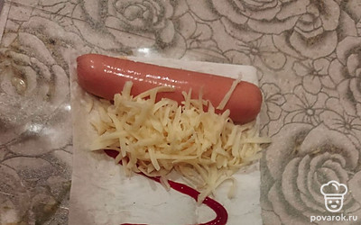 На край лаваша к сосиске добавьте тертый сыр.