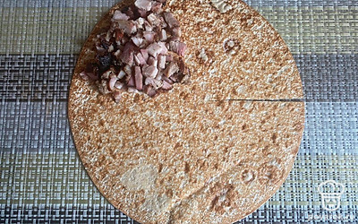 Визуально разделите лавашный круг на 4 части. Сделайте 1 боковой надрез до центра. На одну из частей выложите мелко нарезанную копченую свинину.