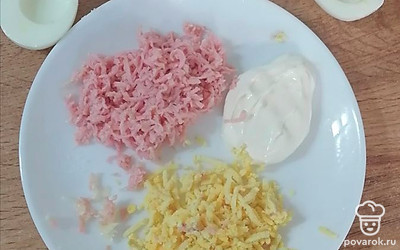 Разрежьте яйца вдоль так, чтобы белок остался с ровными краями. Выньте желток и используйте его в начинке. Сосиску, чеснок и желток натрите на мелкой терке, смешайте со сметаной.