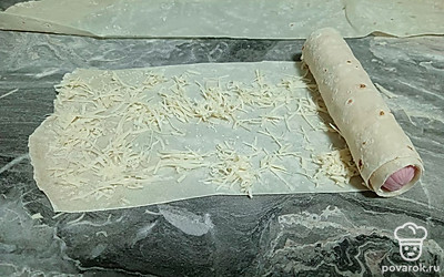 На полоску лаваша выложите тертый сыр, сосиску выложите с краю. И сверните в плотный рулетик.