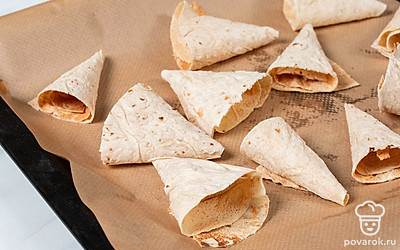 Сложите лаваш вчетверо. Положите на него тарелку, поставленную дном вверх. Ножом обведите контур тарелки, чтобы получились одинаковые круглые листы лаваша. Каждый круг разрежьте пополам и сверните в виде кулечков. Смочите края теста водой, чтобы зафиксировать эти кулечки. Выложите на противень и отправьте в разогретую до 180℃ духовку на 5 минут подсушиться.