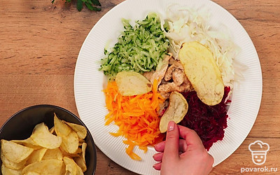 Приготовьте салат. Нарежьте курицу тонкими полосками. Разогрейте сковороду с оливковым маслом, выложите туда курицу. Посолите, поперчите и обжаривайте примерно 15-20 минут. Натрите на терке морковь, огурец и свеклу. Из огурца отожмите сок. Возьмите большую плоскую тарелку. В центр выложите обжаренное куриное филе. Вокруг небольшими порциями выложите все овощи. В конце добавьте чипсы, положив их сверху.